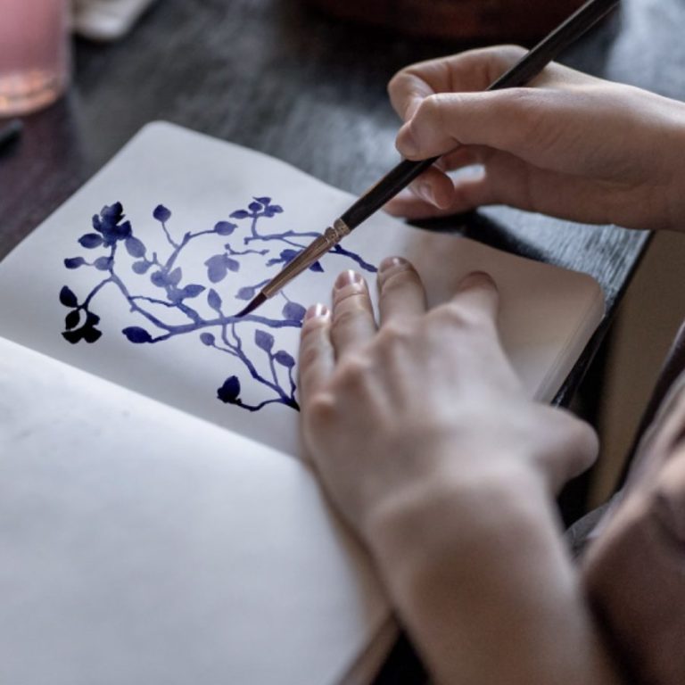 Hände halten einen Pinsel und malen blauen Blumen in ein Skizzenbuch.